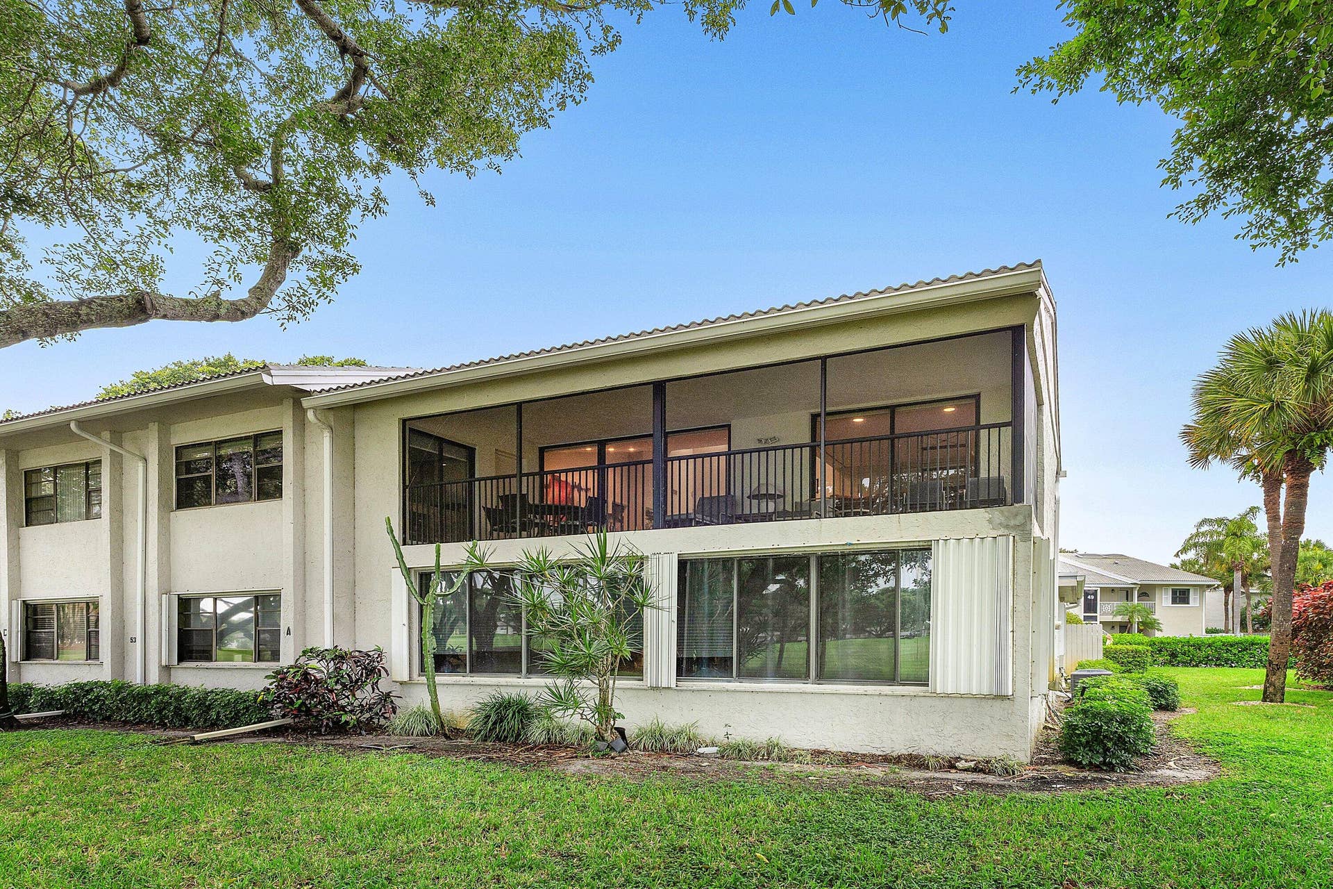 53 EASTGATE Drive, #B, Boynton Beach, FL 33436 Two-story exterior featuring a screened-in second-level balcony with metal railing, floor-to-ceiling windows on the ground level, and mature landscaping