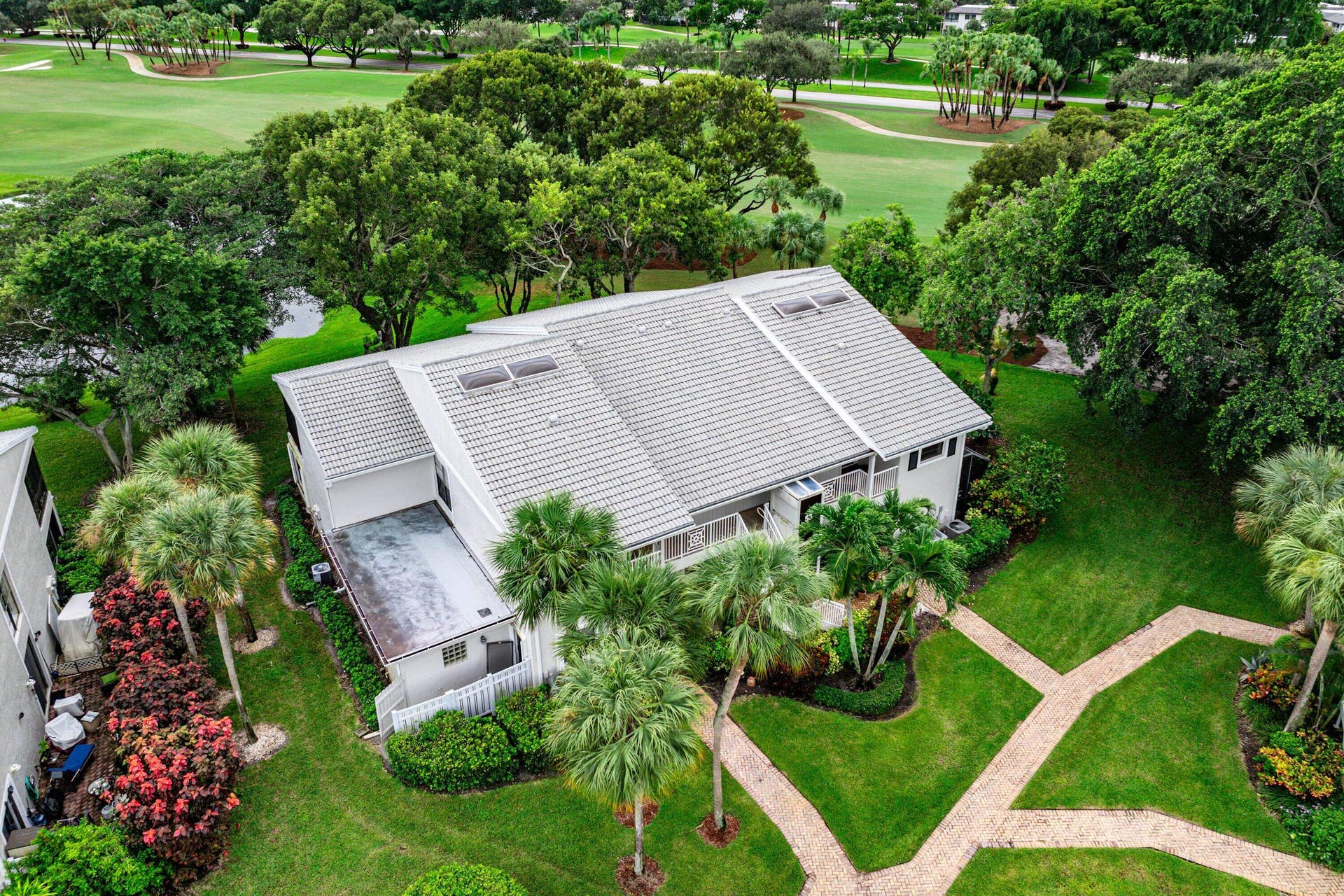 53 EASTGATE Drive, #B, Boynton Beach, FL 33436 Exterior aerial featuring a multi-pitched roof with integrated skylights, a light-toned facade, mature palm trees, and manicured landscaping with paver walkways