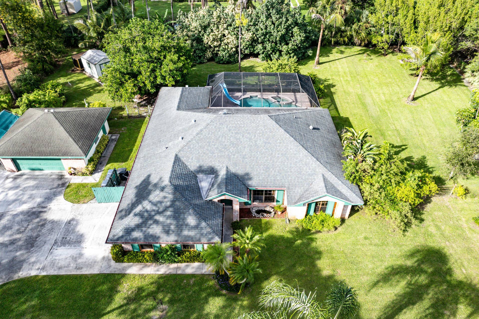 13677 Citrus Grove Boulevard, West Palm Beach, FL 33412 Aerial perspective featuring a multi-gabled shingle roof, expansive manicured grounds, a screened-in pool enclosure with an integrated water slide, a detached garage, and an extensive concrete driveway