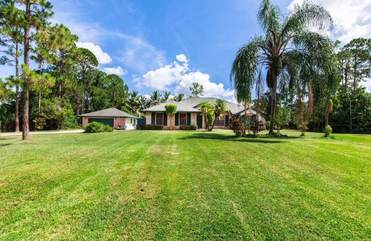 13677 Citrus Grove Boulevard, West Palm Beach, FL 33412 Single-story residence featuring a brick facade, detached 2.5 car garage, expansive lawn, and mature palm trees