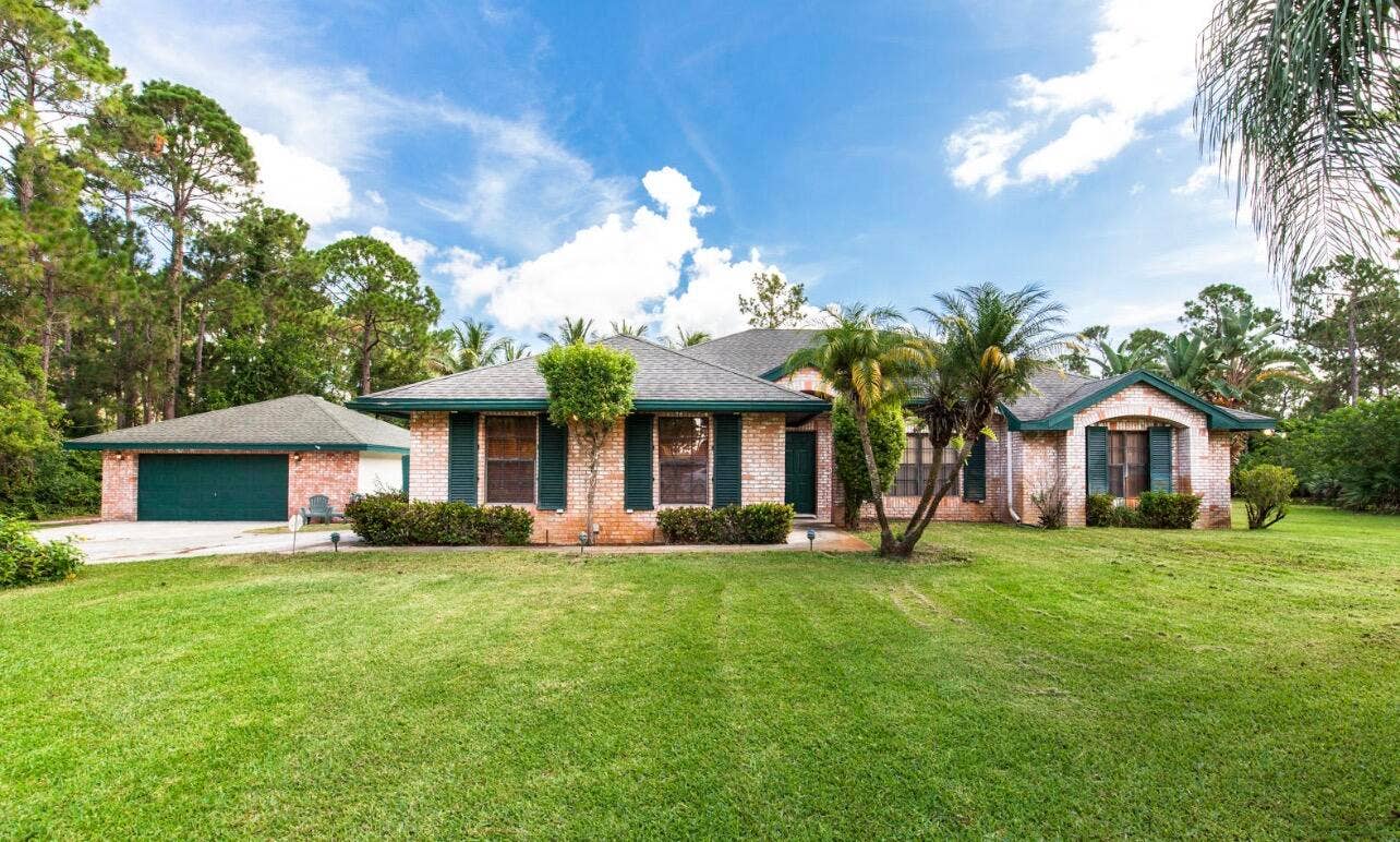 13677 Citrus Grove Boulevard, West Palm Beach, FL 33412 Single-story residence featuring a brick facade, detached 2.5 car garage, expansive lawn, and mature palm trees