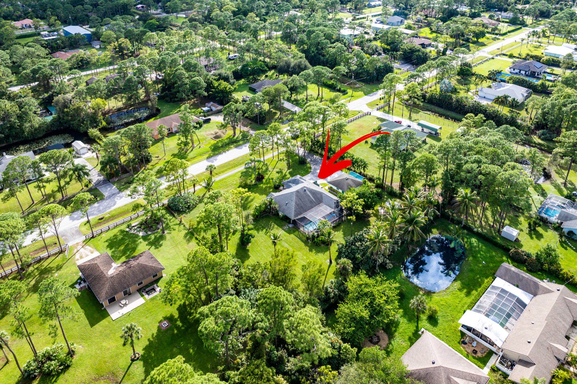 13677 Citrus Grove Boulevard, West Palm Beach, FL 33412 Aerial perspective of a detached residence featuring a multi-slope roof, an attached garage, a screen-enclosed pool, and a private driveway situated on a wooded lot