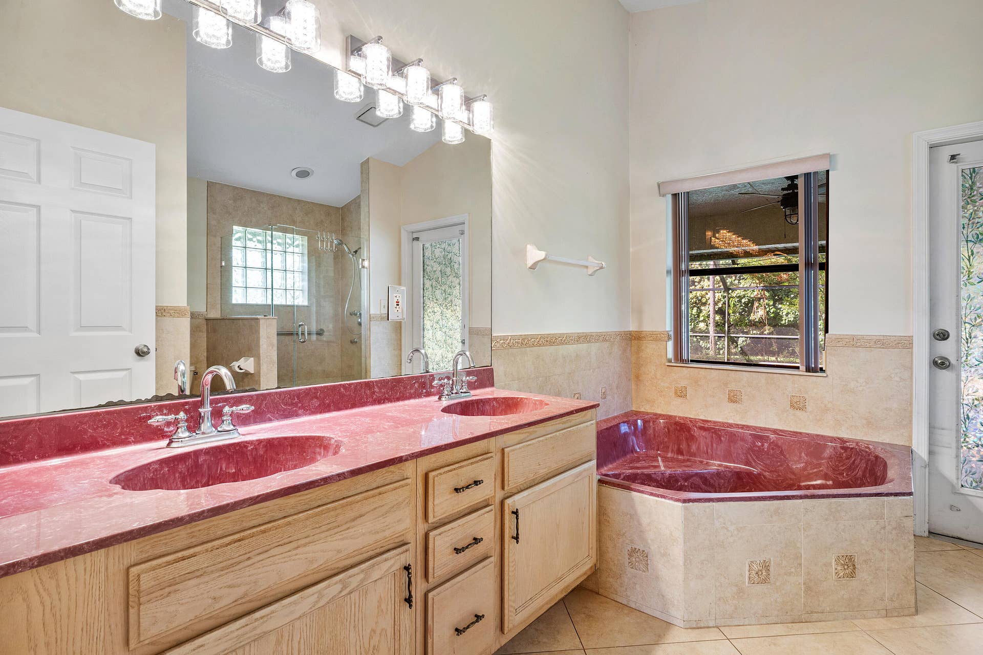 13677 Citrus Grove Boulevard, West Palm Beach, FL 33412 Primary bathroom ensuite number one double vanity featuring a wide countertop with dual integrated sinks, light wood cabinetry, a large wall-mounted mirror, multi-bulb overhead lighting, and a corner soaking tub finished with decorative stone tile accents