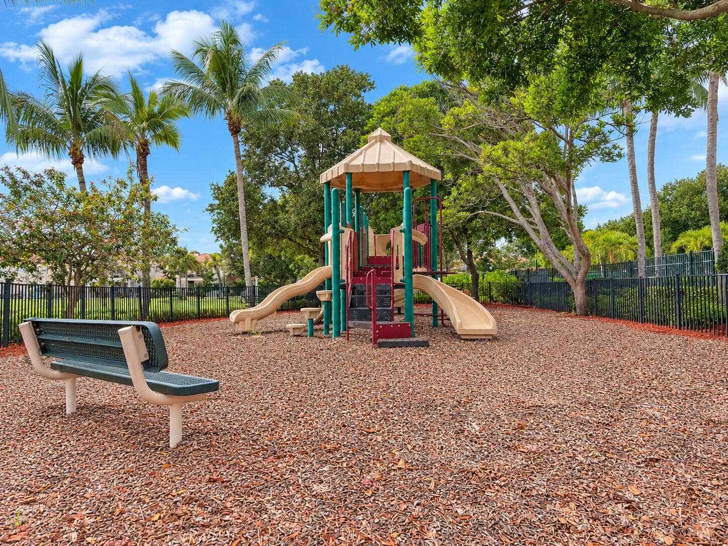 762 GAZETTA Way, West Palm Beach, FL 33413 Outdoor recreation area featuring a multi-level play structure with dual slides, wood-chip ground cover, a metal-frame bench, and perimeter fencing shaded by mature palm and deciduous trees