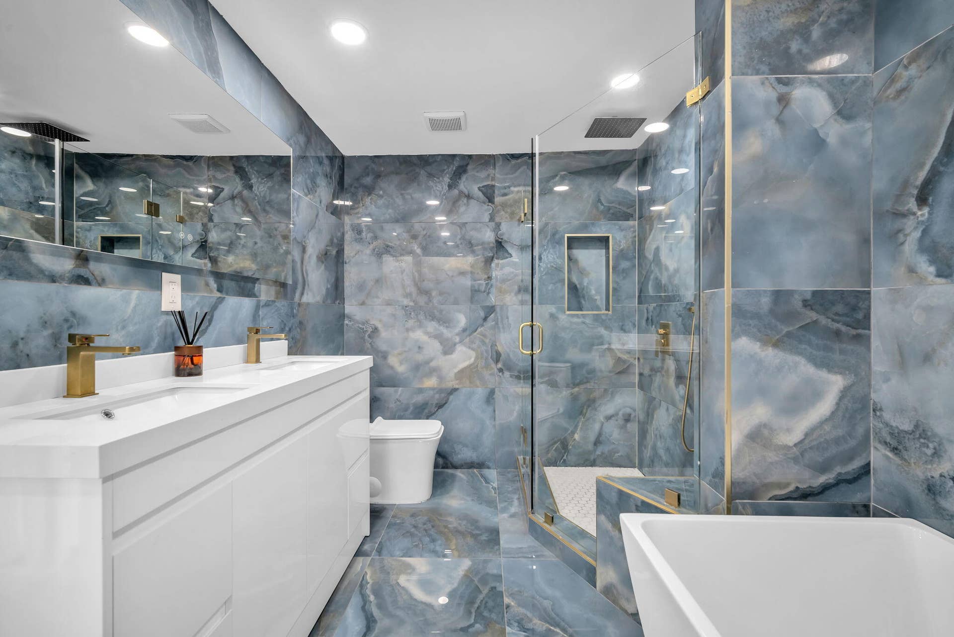 4162 SW 65th Avenue, #84, Davie, FL 33314 Bathroom featuring full-height blue decorative stone tiling, white double-vanity cabinet with integrated sinks, brass-finish fixtures, glass-enclosed walk-in shower with built-in recessed shelving, and a white freestanding soaking tub