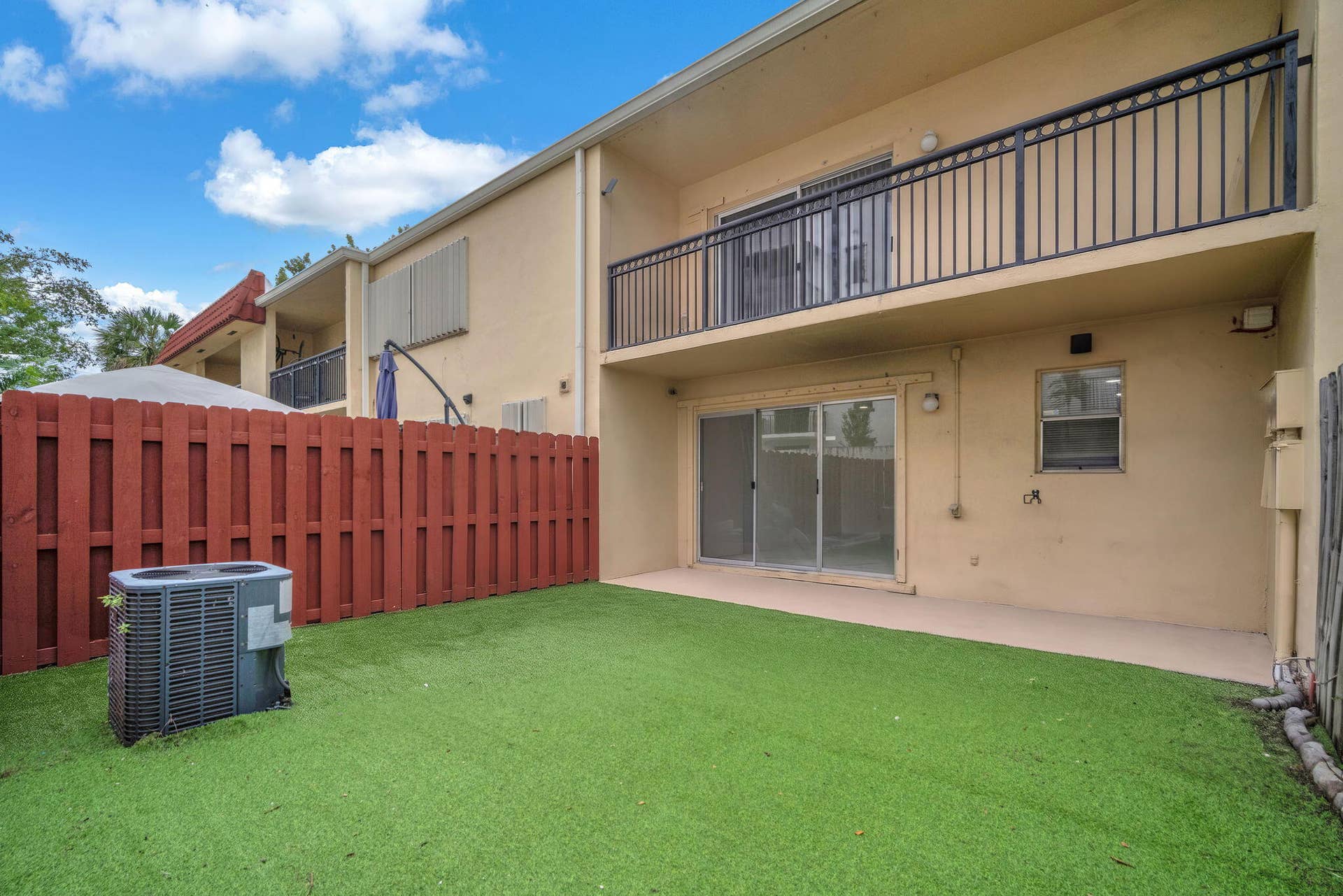 4162 SW 65th Avenue, #84, Davie, FL 33314 Private backyard with synthetic turf, enclosed by a wood-slat privacy fence, featuring a concrete patio, sliding glass doors, and a second-story balcony with metal railings