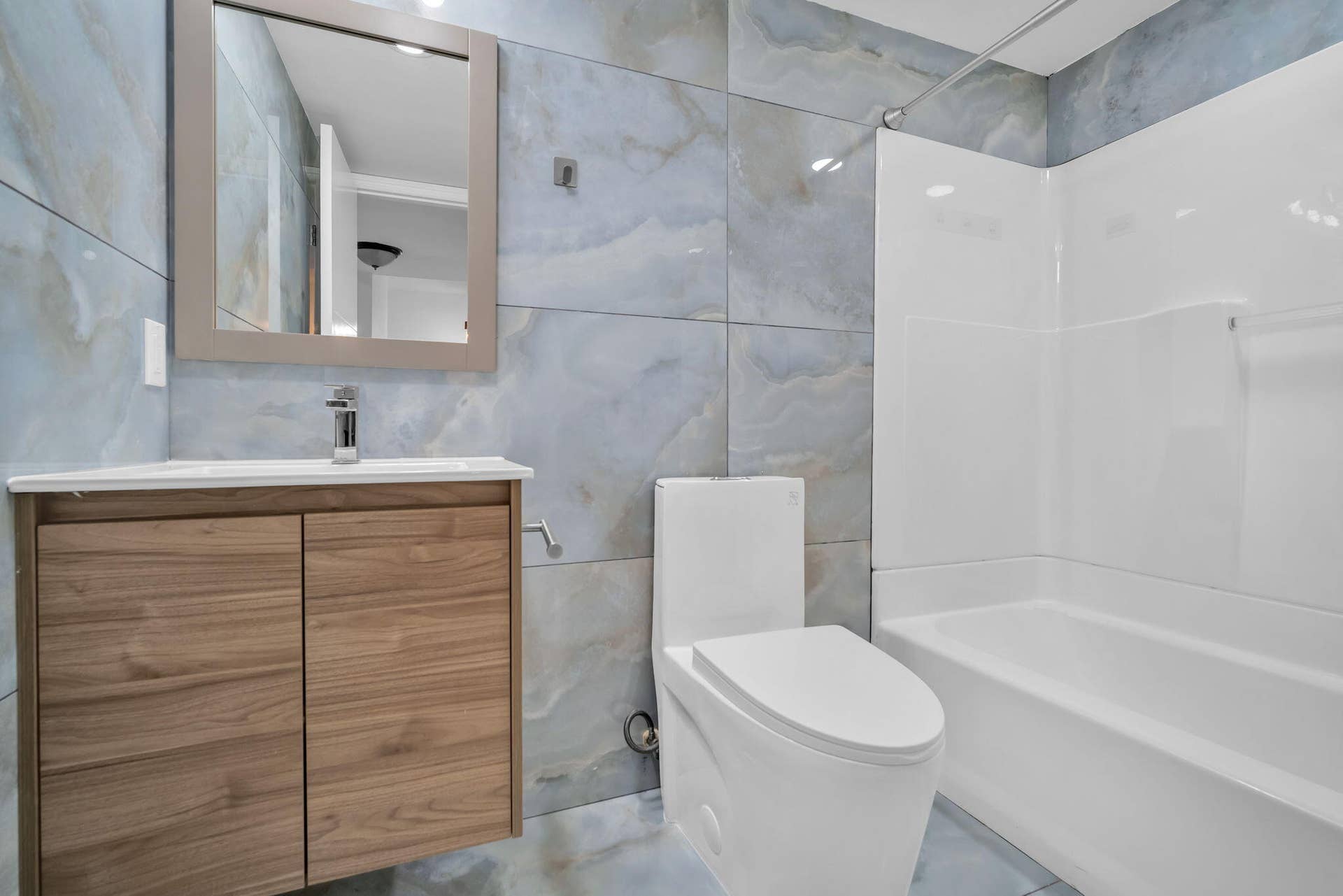 4162 SW 65th Avenue, #84, Davie, FL 33314 Full-height wall tiling with large-format blue-toned decorative stone-finish panels, wall-mounted wood-finish vanity with a rectangular basin, polished chrome faucet, and white acrylic tub and shower surround