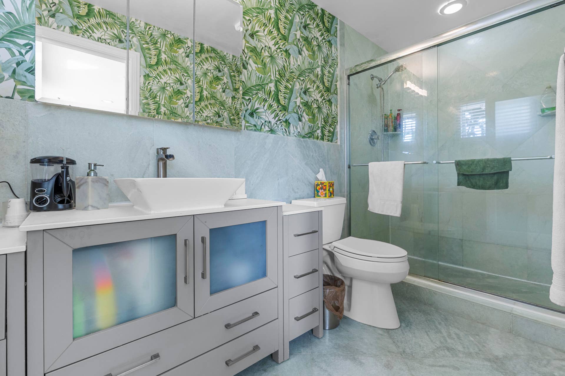 2317 NW 6th Avenue, Wilton Manors, FL 33311 Bathroom featuring a grey vanity with frosted glass cabinet doors, a rectangular vessel sink, stone-finish wall tile, a walk-in shower with a glass enclosure, and patterned wallpaper