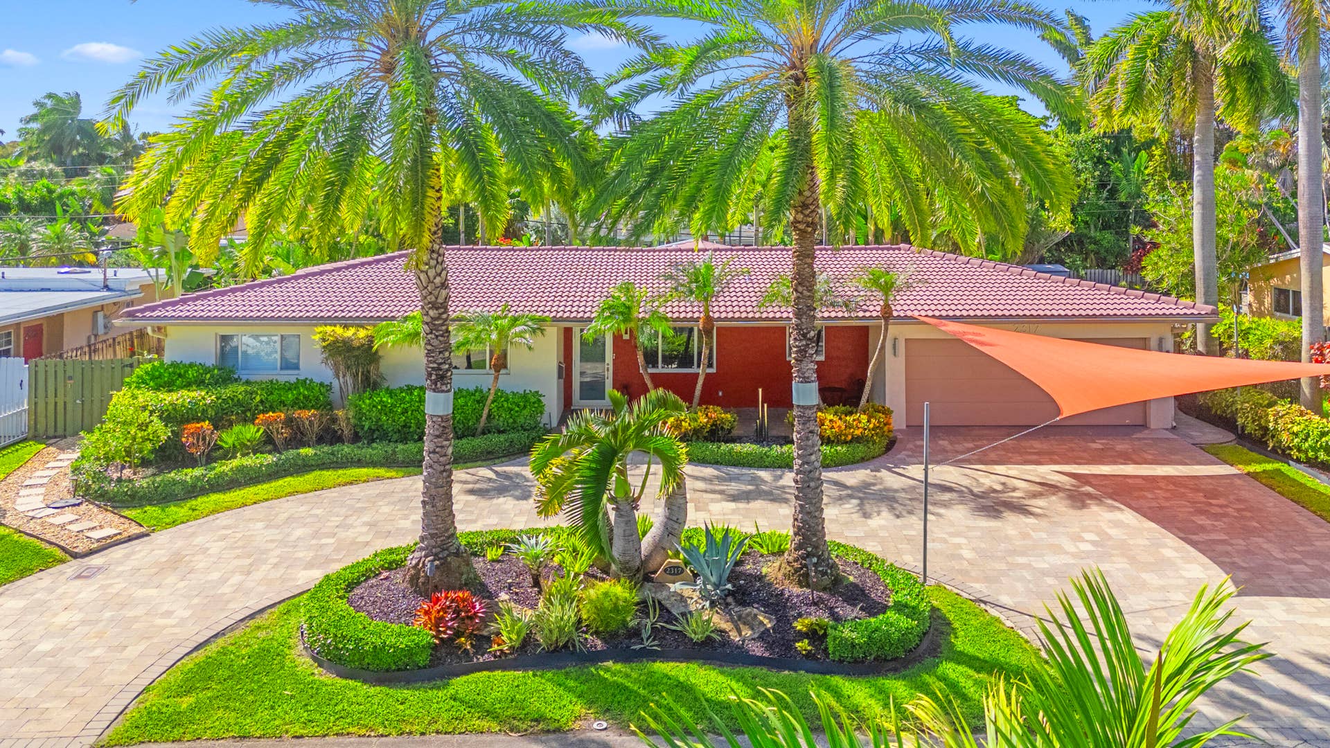 2317 NW 6th Avenue, Wilton Manors, FL 33311 Single-story residence featuring a tile roof, paved circular driveway, mature palm trees, professional landscaping, and a two-car garage