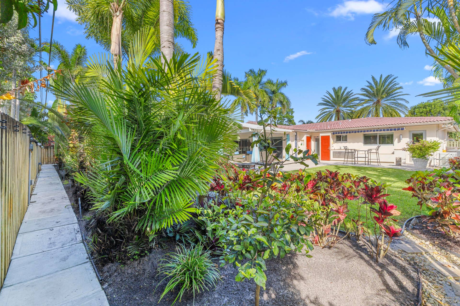 2317 NW 6th Avenue, Wilton Manors, FL 33311 Rear exterior featuring a concrete walkway, mature palm trees, diverse tropical landscaping, and a fenced perimeter
