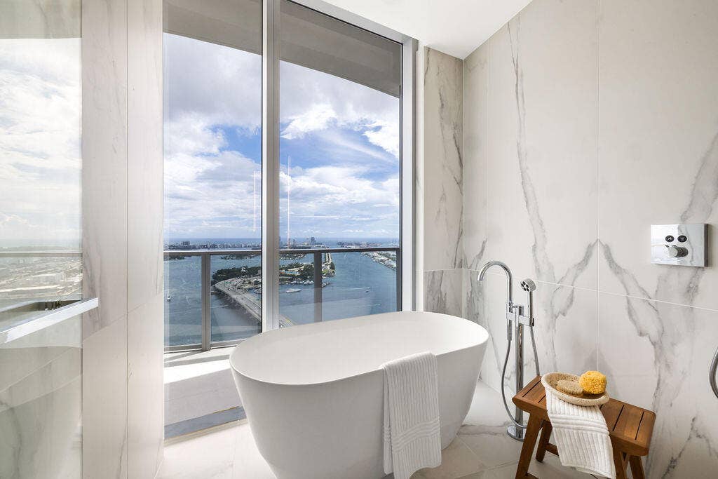 Freestanding soaking tub with polished chrome floor-mounted faucet, floor-to-ceiling porcelain wall tile, expansive window wall, and integrated balcony with metal railing