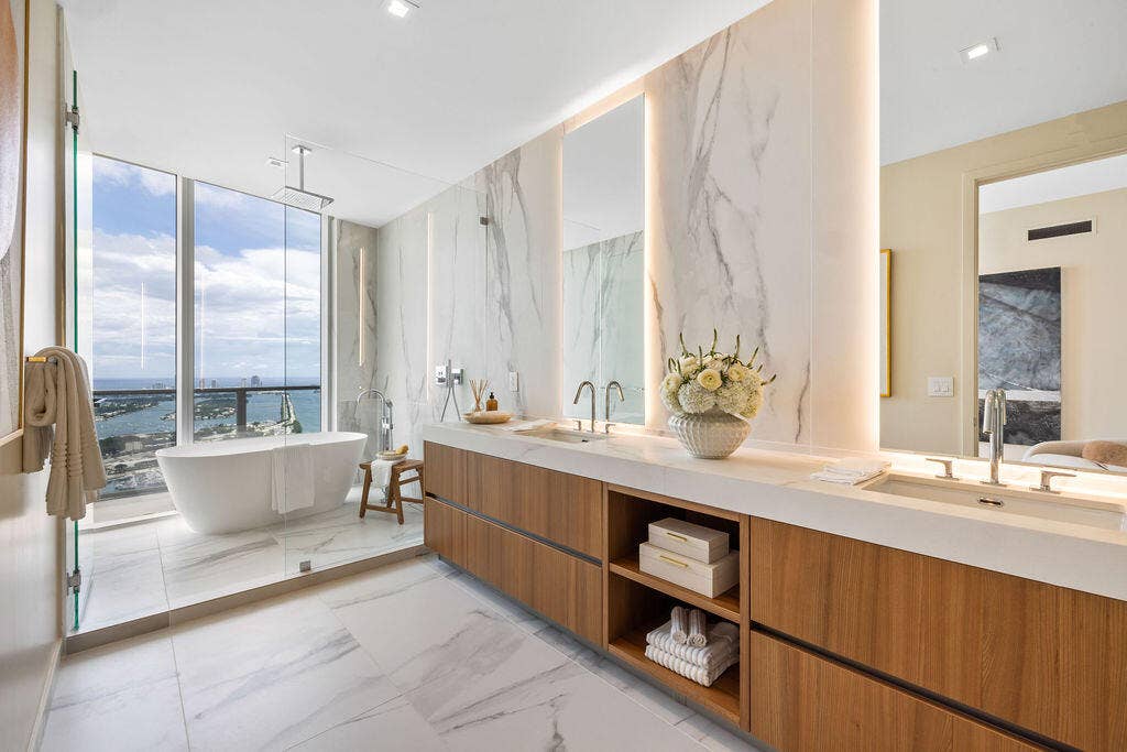 Bathroom featuring floor-to-ceiling stone wall cladding, a floating wood-finish dual vanity with integrated countertop basins, a deep soaking tub, and expansive floor-to-ceiling windows