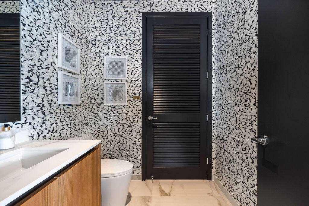 High-contrast patterned wall tile, vanity with a rectangular sink, light-toned countertop, black louvered door, and large-format neutral tile flooring