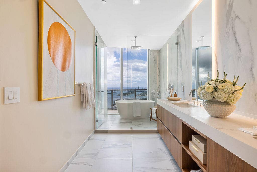 Floating vanity with wood-finish cabinetry, solid-surface countertop, full-height stone-finish wall cladding, and a freestanding soaking tub enclosed in a glass shower space