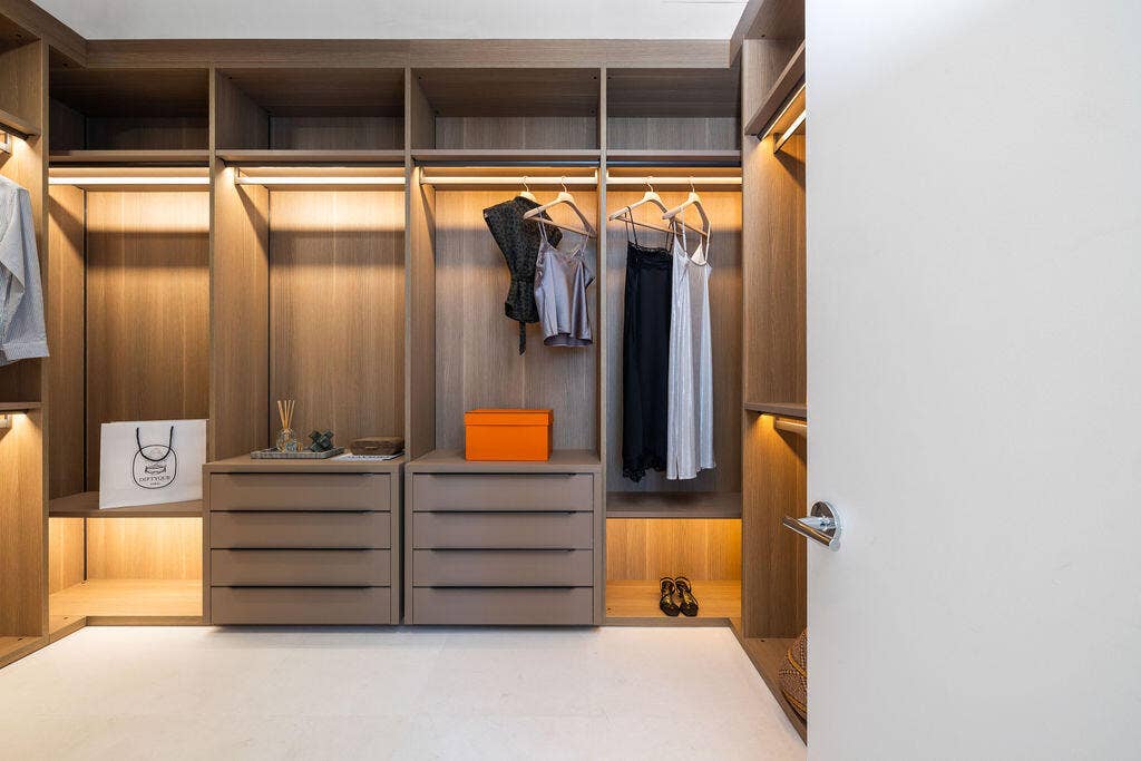 Custom reach-in closet featuring wood-finish cabinetry, built-in dresser drawers with matte hardware, integrated LED task lighting, and open shelving