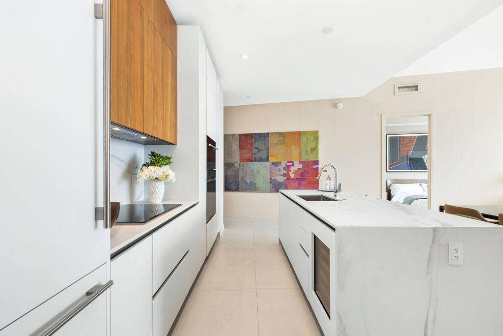 Kitchen featuring white flat-panel cabinetry, wood-finish upper cabinets, stone-surfaced waterfall island, built-in wall oven, and integrated wine cooler