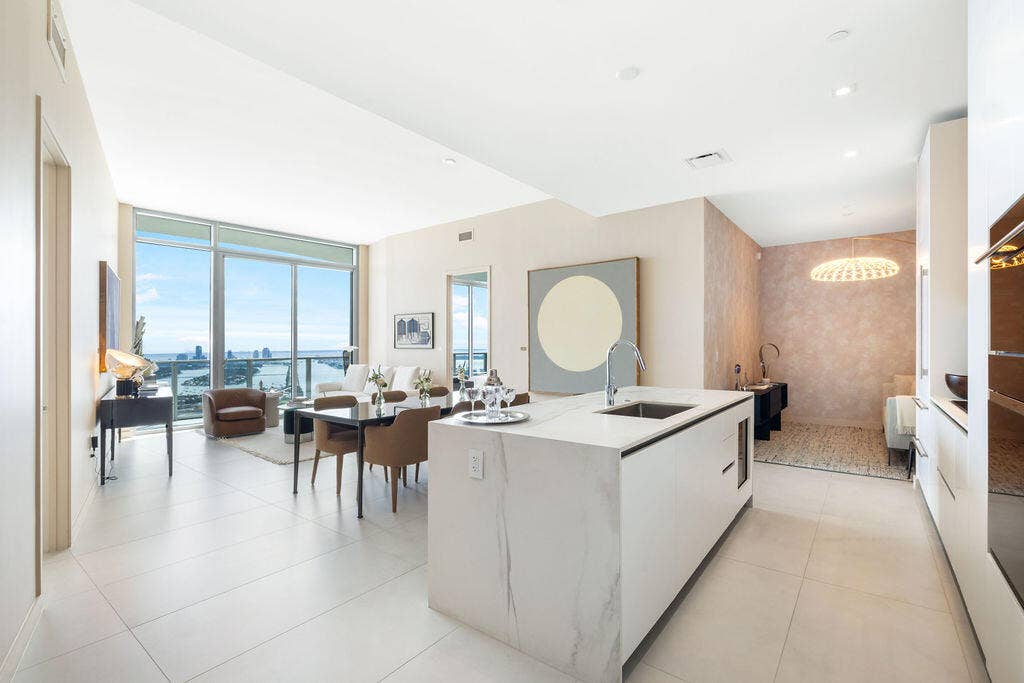 Open-concept interior featuring an oversized waterfall-edge stone island, floor-to-ceiling glass windows, high ceilings with recessed lighting, and expansive light-toned tile flooring