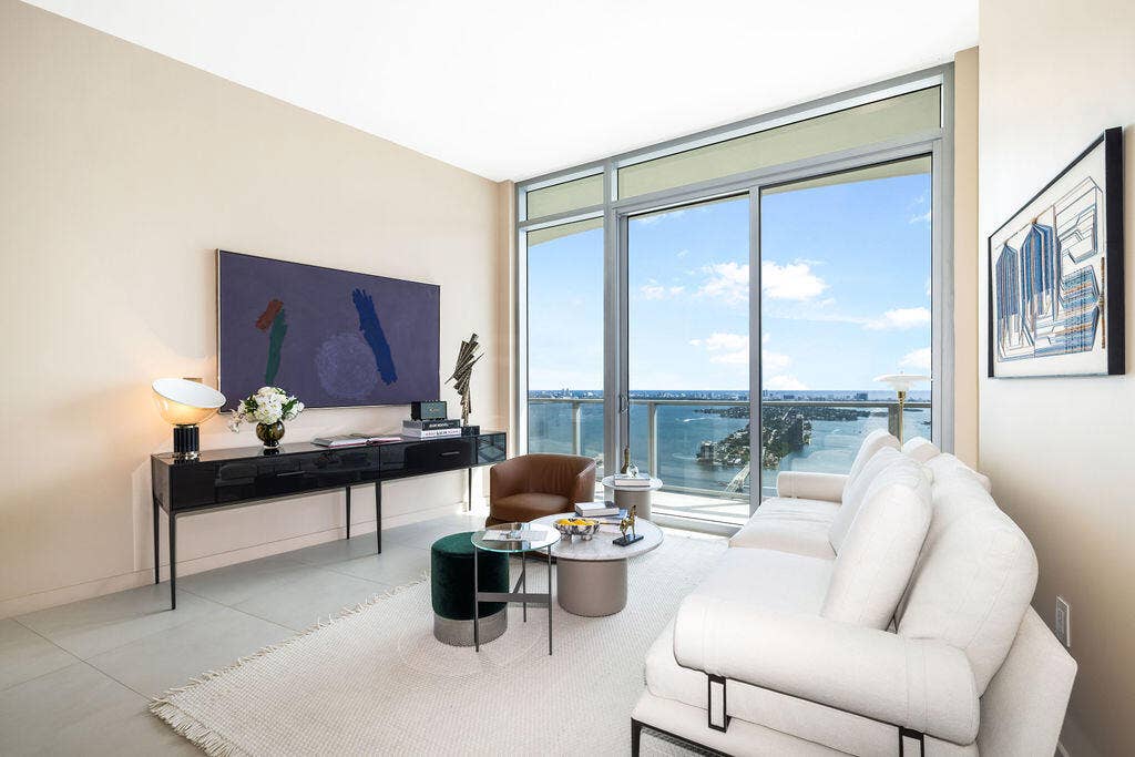 Floor-to-ceiling windows, sliding glass door, private balcony, neutral large-format tile flooring, and water backdrop