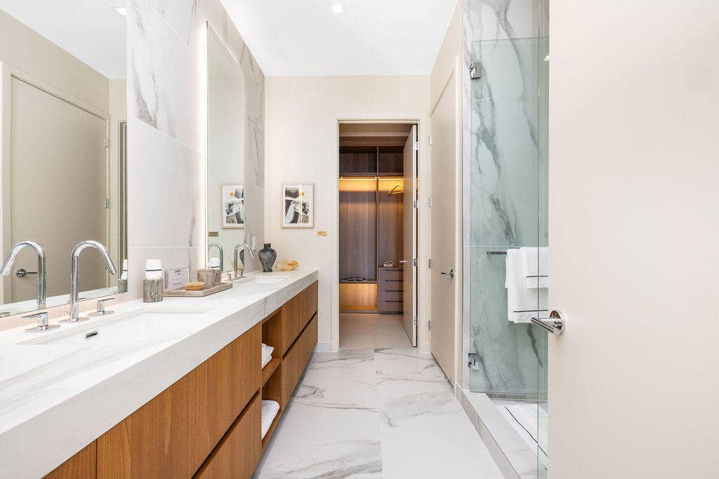 Dual-sink vanity with wood-finish cabinetry, stone-surfaced countertop, full-height backlit mirrors, and glass-enclosed shower with large-format tile wall cladding