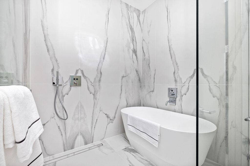 Freestanding soaking tub featuring a wall-mounted faucet, floor-to-ceiling large-format stone-finish tile surround, handheld shower fixture, and glass shower enclosure