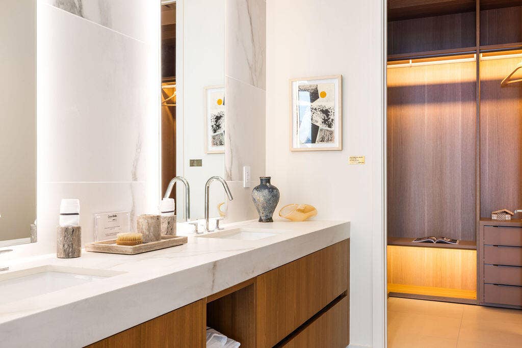 Floating vanity with dual rectangular undermount sinks, stone-finish countertops, integrated LED mirror lighting, and custom wood-finish cabinetry including a built-in closet system with integrated drawer storage
