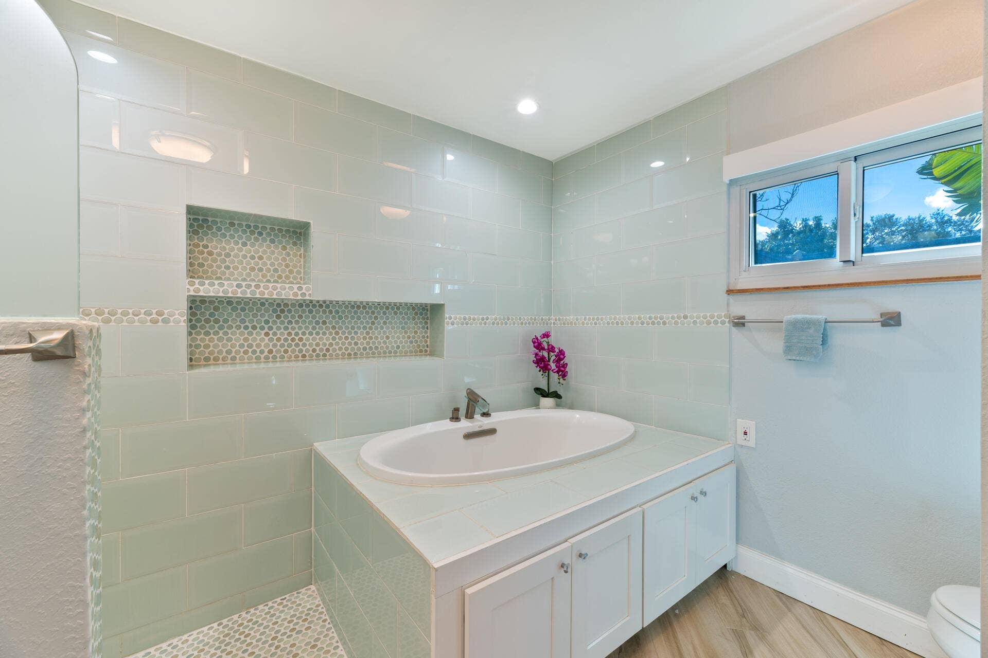 Built-in soaking tub with a tiled surround, recessed wall niche with decorative accent tiling, wood-finish flooring, and recessed lighting
