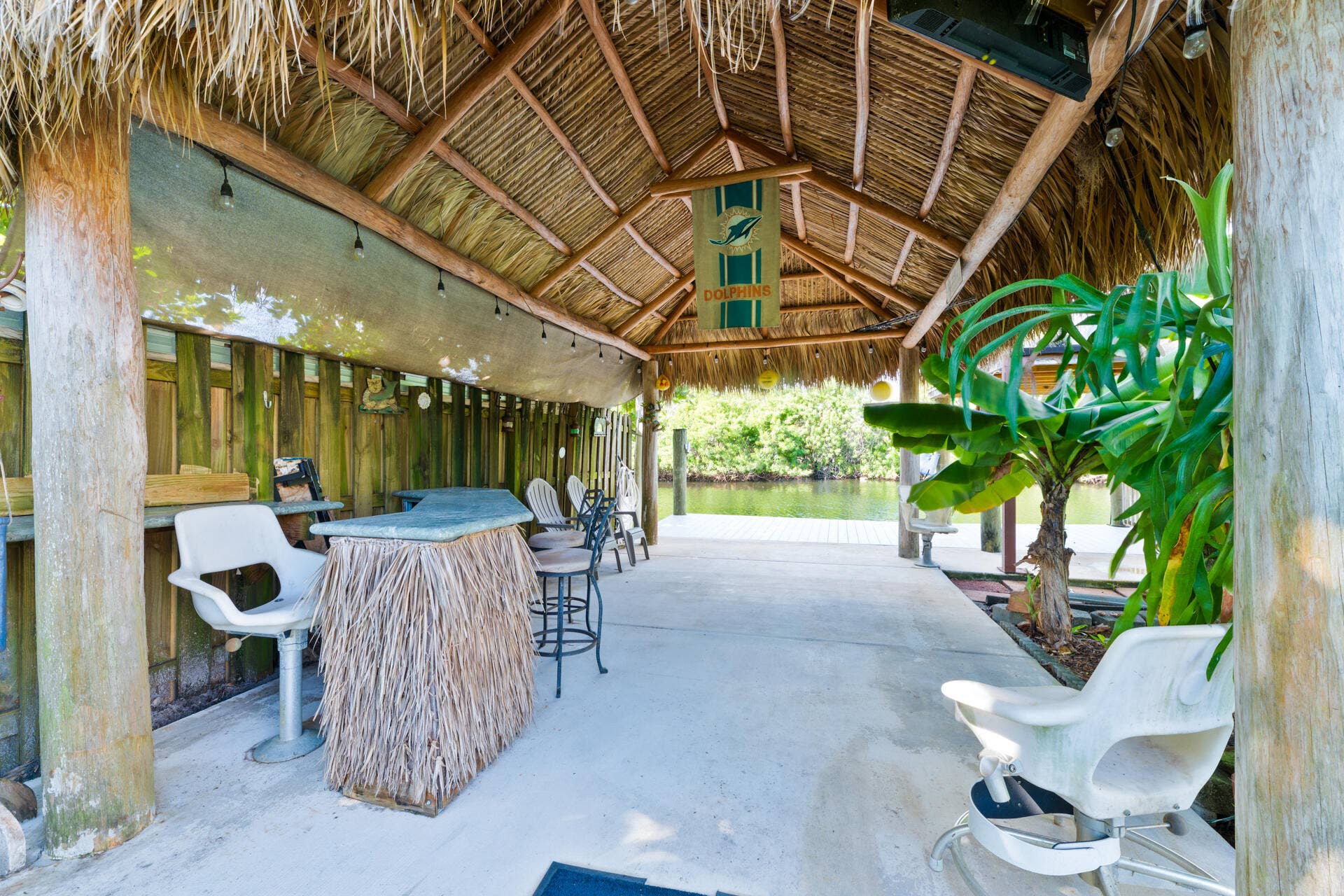 Covered outdoor tiki bar featuring a thatched roof, exposed timber support posts, concrete flooring, and stand alone bar