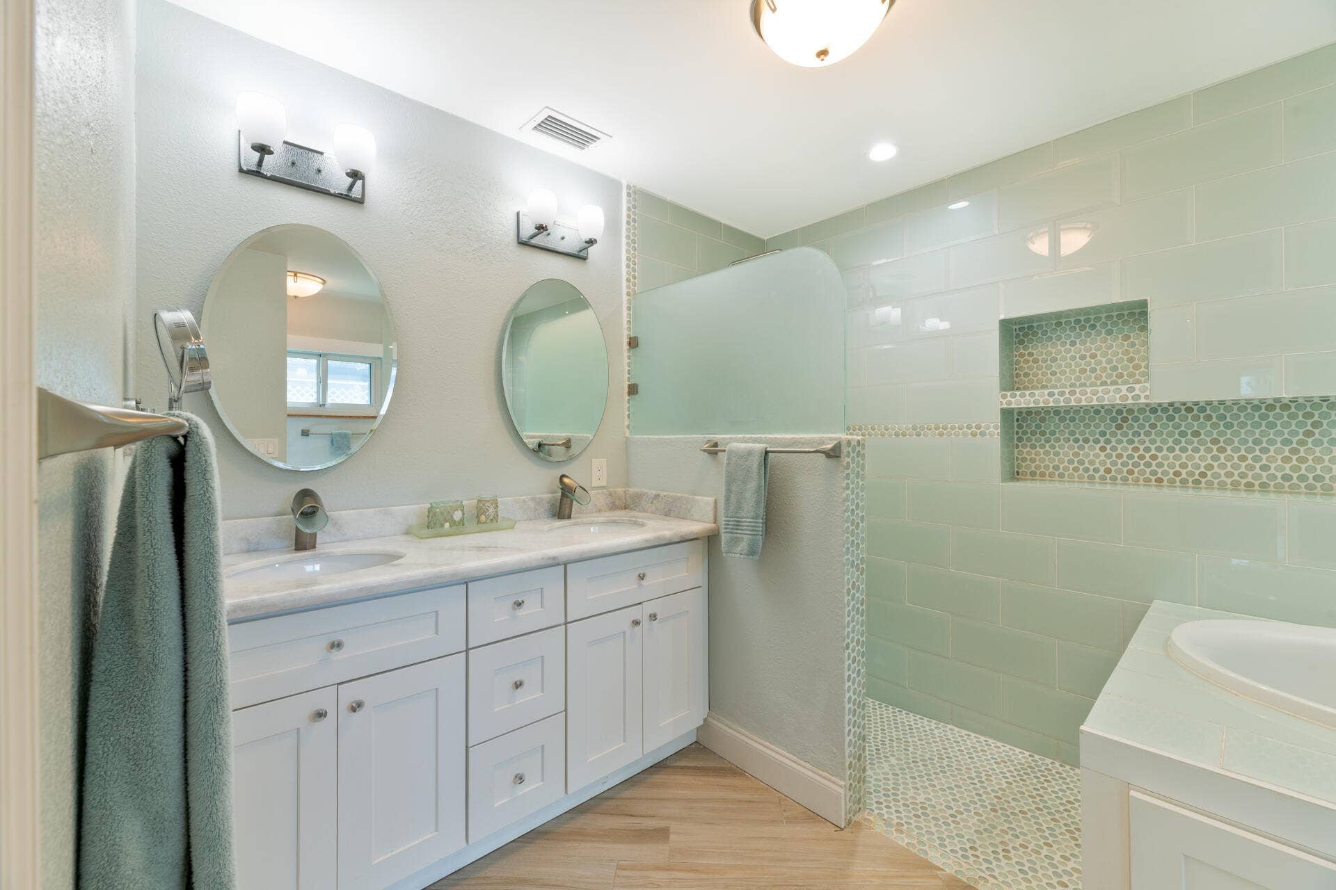 Dual-sink vanity with stone countertop, walk-in shower with tile surround and integrated shelving, wood-finish flooring, and dual oval wall mirrors