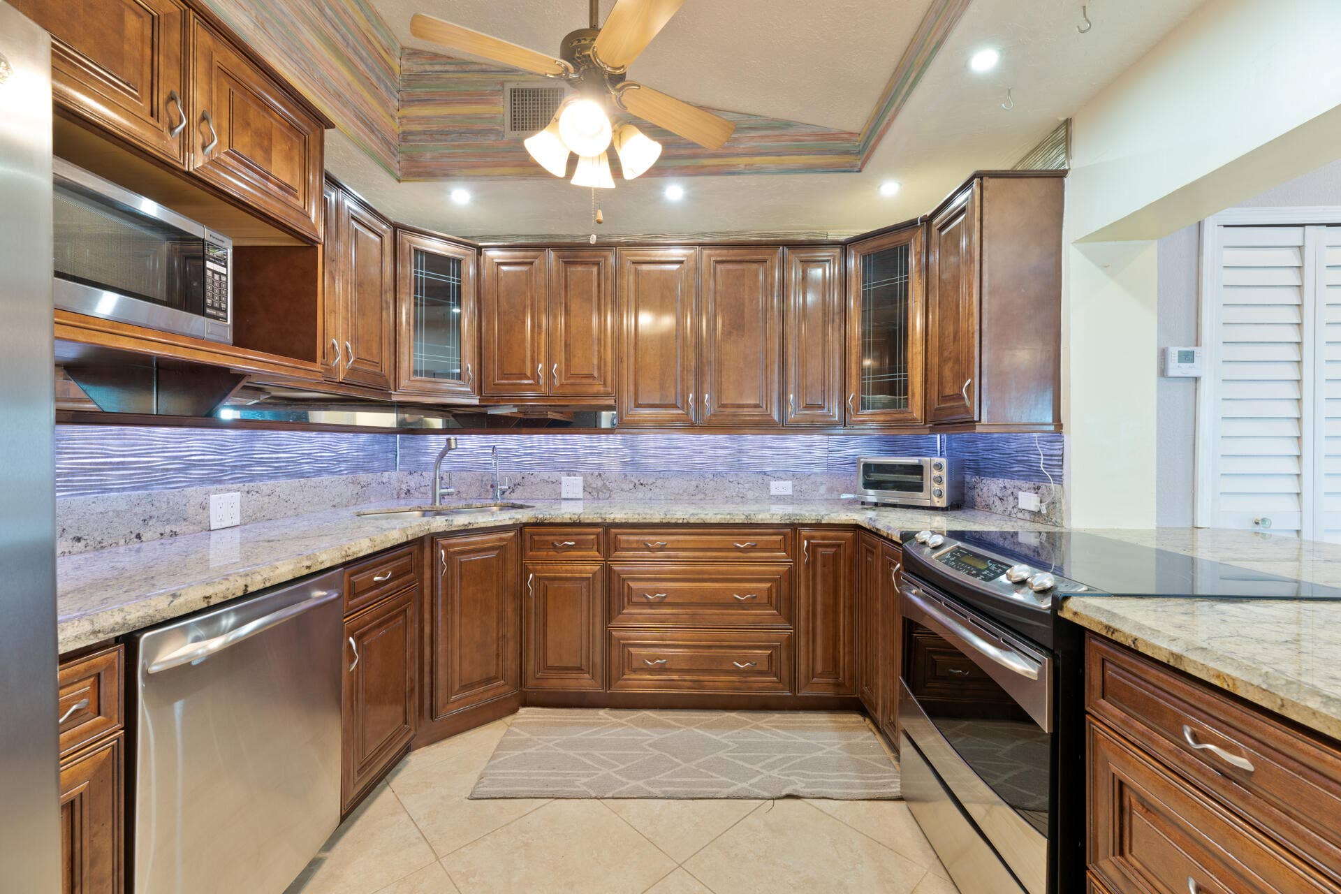 Custom wood cabinetry featuring glass-front display panels, stone countertops, textured tile backsplash, stainless steel appliances, and tray ceiling with recessed lighting