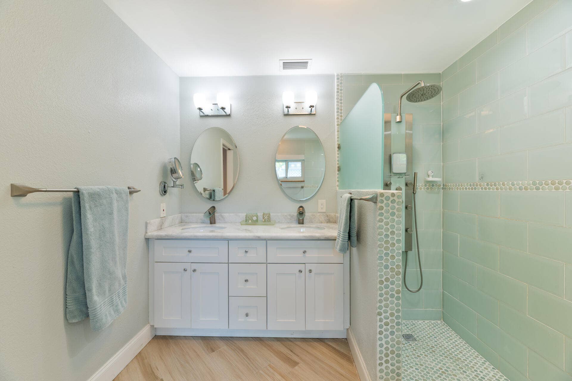 Dual-sink vanity with white cabinetry and stone countertop, dual oval wall mirrors, walk-in shower featuring glass tile surround, and wood-finish flooring