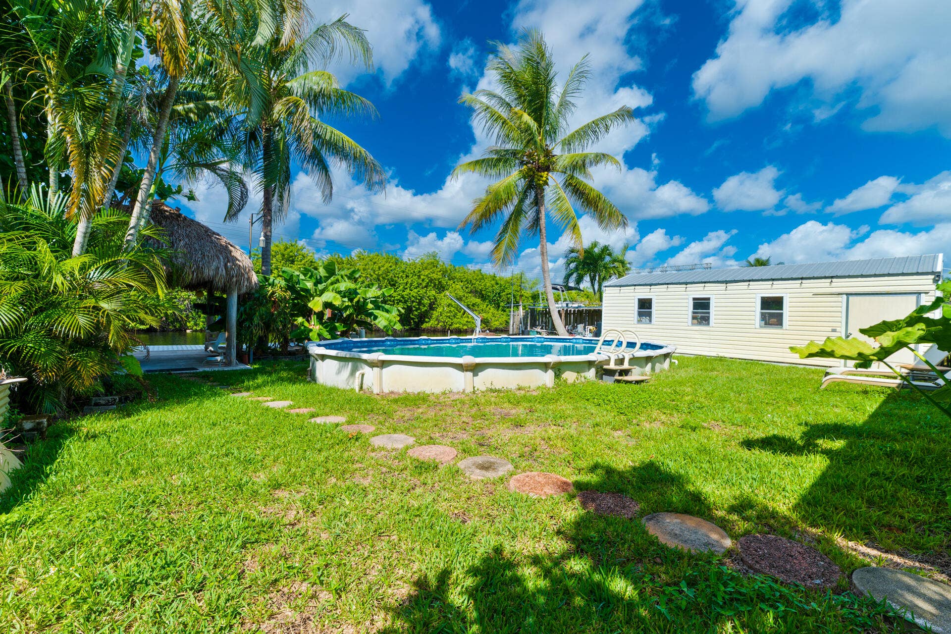 Backyard featuring an above-ground pool, stone paver pathway, detached storage/workshop structure, and mature palm trees