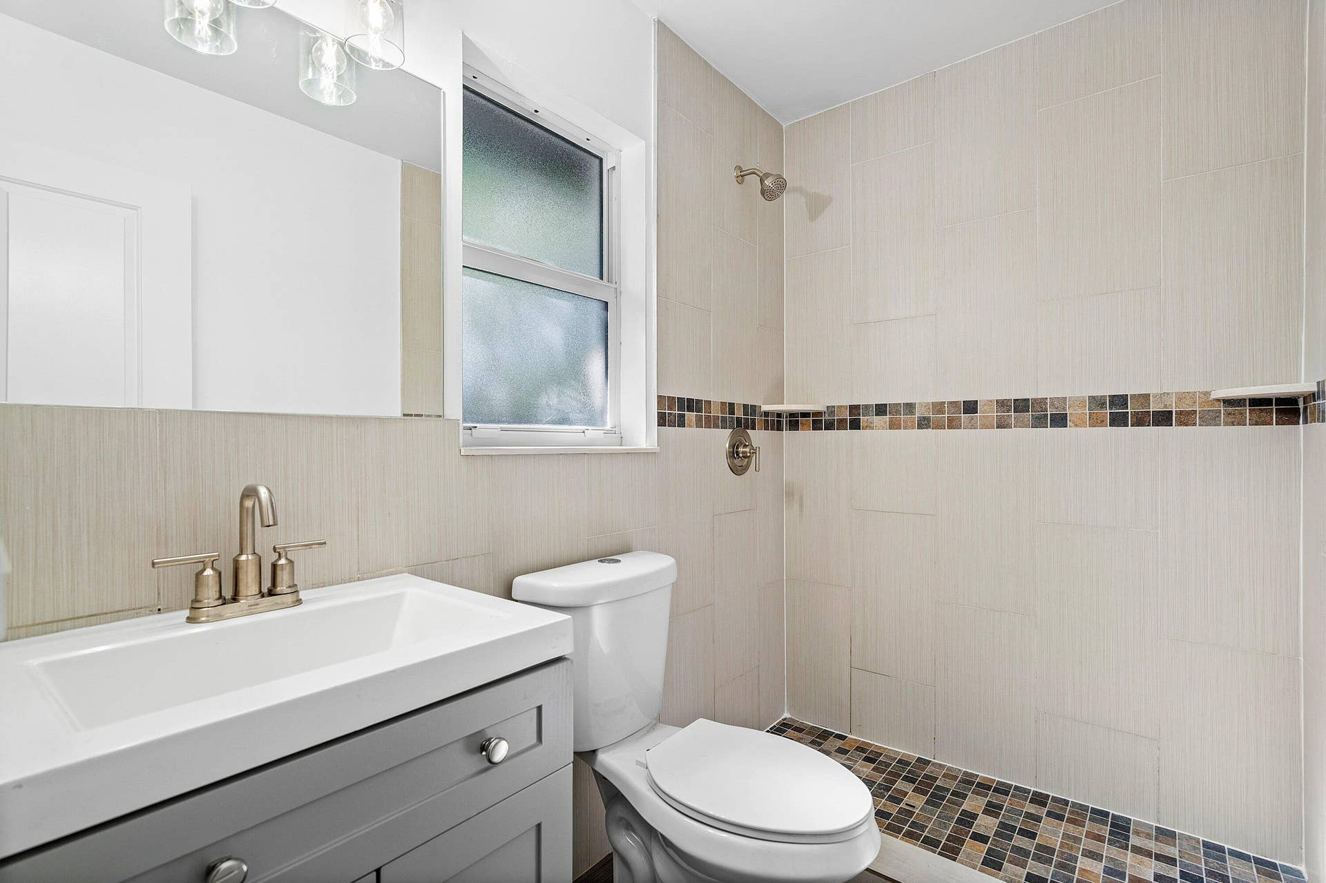 1211 NE 2nd Avenue, Fort Lauderdale, FL 33304 Bathroom featuring a gray vanity with an integrated sink, brushed-metal hardware, a wall-mounted mirror, and a walk-in shower with neutral tile and decorative mosaic accents