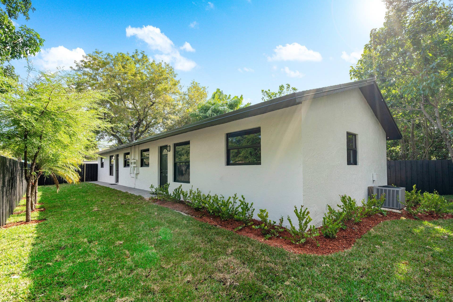 1211 NE 2nd Avenue, Fort Lauderdale, FL 33304 Stucco exterior featuring dark-trimmed windows, a multi-paneled door, manicured landscaping with mulch beds, and an enclosed fenced yard