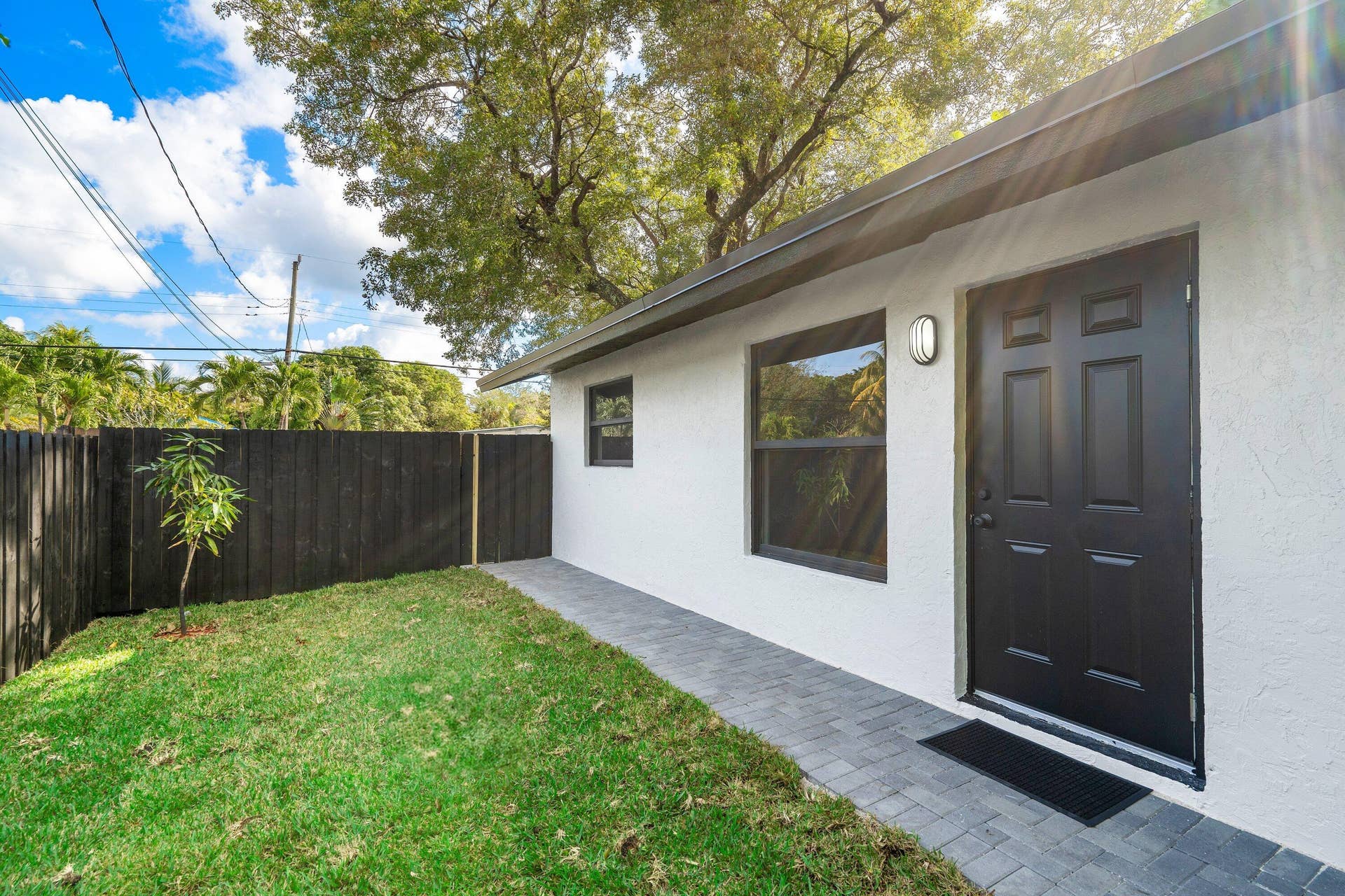1211 NE 2nd Avenue, Fort Lauderdale, FL 33304 Exterior featuring a white stucco facade, a dark-finished panel door, a grey paver walkway, and a fenced yard with mature trees