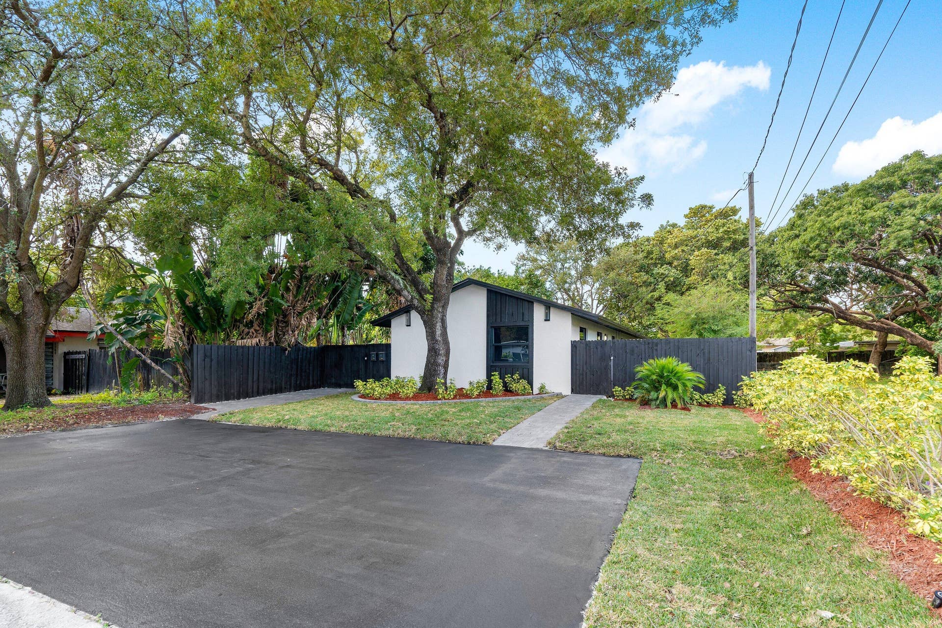 1211 NE 2nd Avenue, Fort Lauderdale, FL 33304 Exterior featuring a white facade with black architectural accents, asphalt driveway, mature trees, and enclosed perimeter fencing