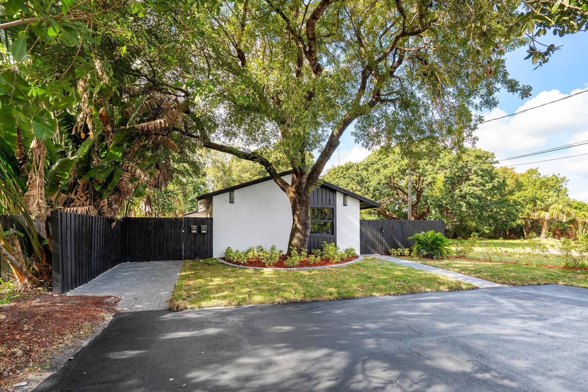 1211 NE 2nd Avenue, Fort Lauderdale, FL 33304 Exterior facade featuring white stucco walls, vertical dark-toned siding, mature tree canopy, stone-paver driveway, and solid dark-toned privacy fencing