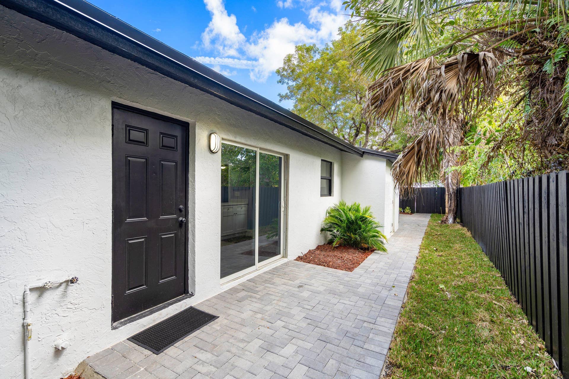 1211 NE 2nd Avenue, Fort Lauderdale, FL 33304 Exterior space featuring white stucco siding, dark-toned panel door, sliding glass door, grey paver patio, and dark-toned perimeter fencing