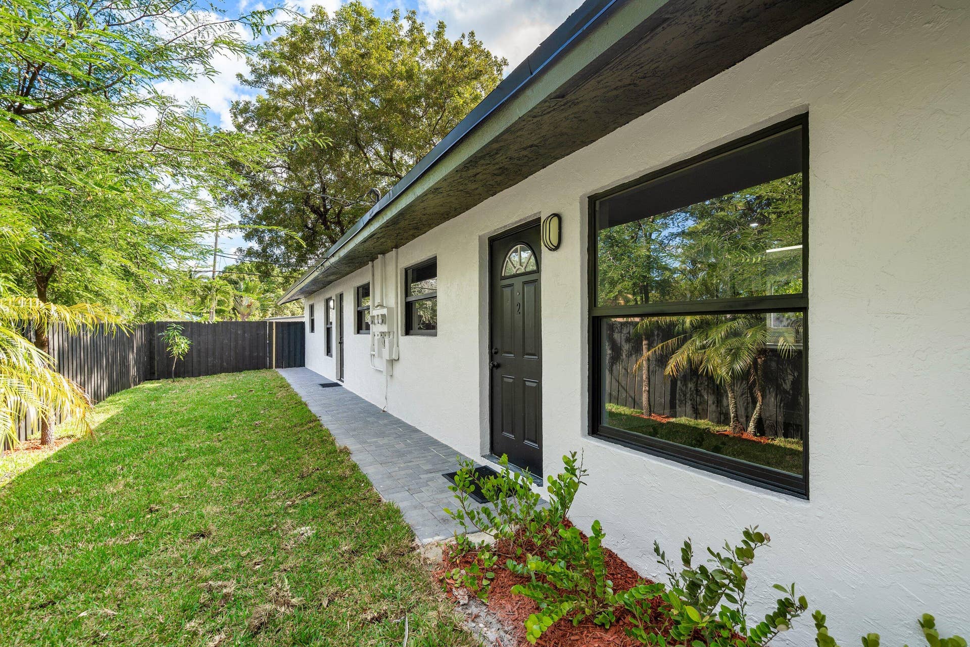 1211 NE 2nd Avenue, Fort Lauderdale, FL 33304 Exterior featuring white stucco siding, dark-toned window frames, paver walkway, and fenced yard space with mature trees