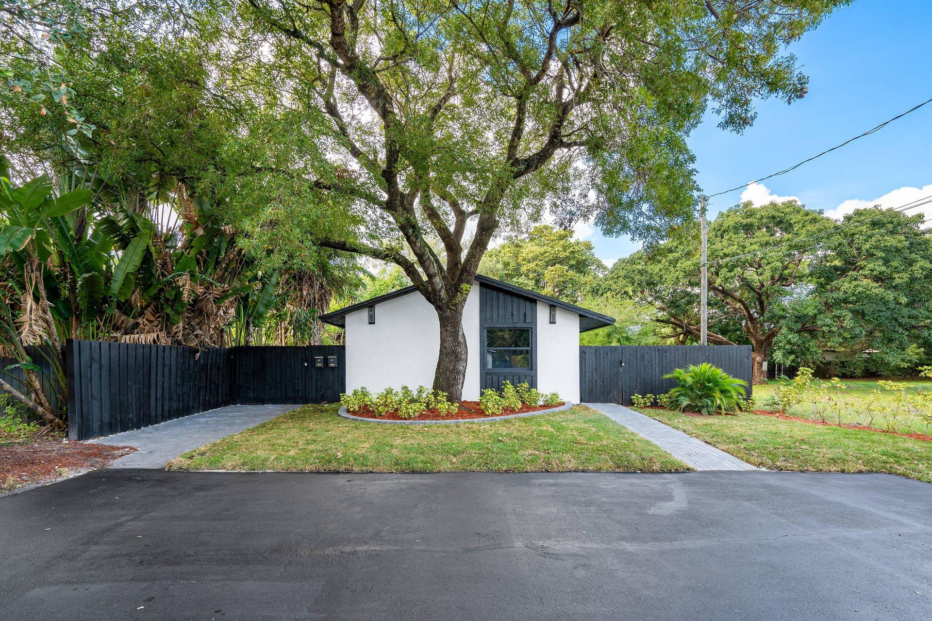 1211 NE 2nd Avenue, Fort Lauderdale, FL 33304 Exterior facade featuring white siding with dark-toned trim, asphalt-paved driveway, decorative landscaping with a central mature tree, and dark wood-style privacy fencing