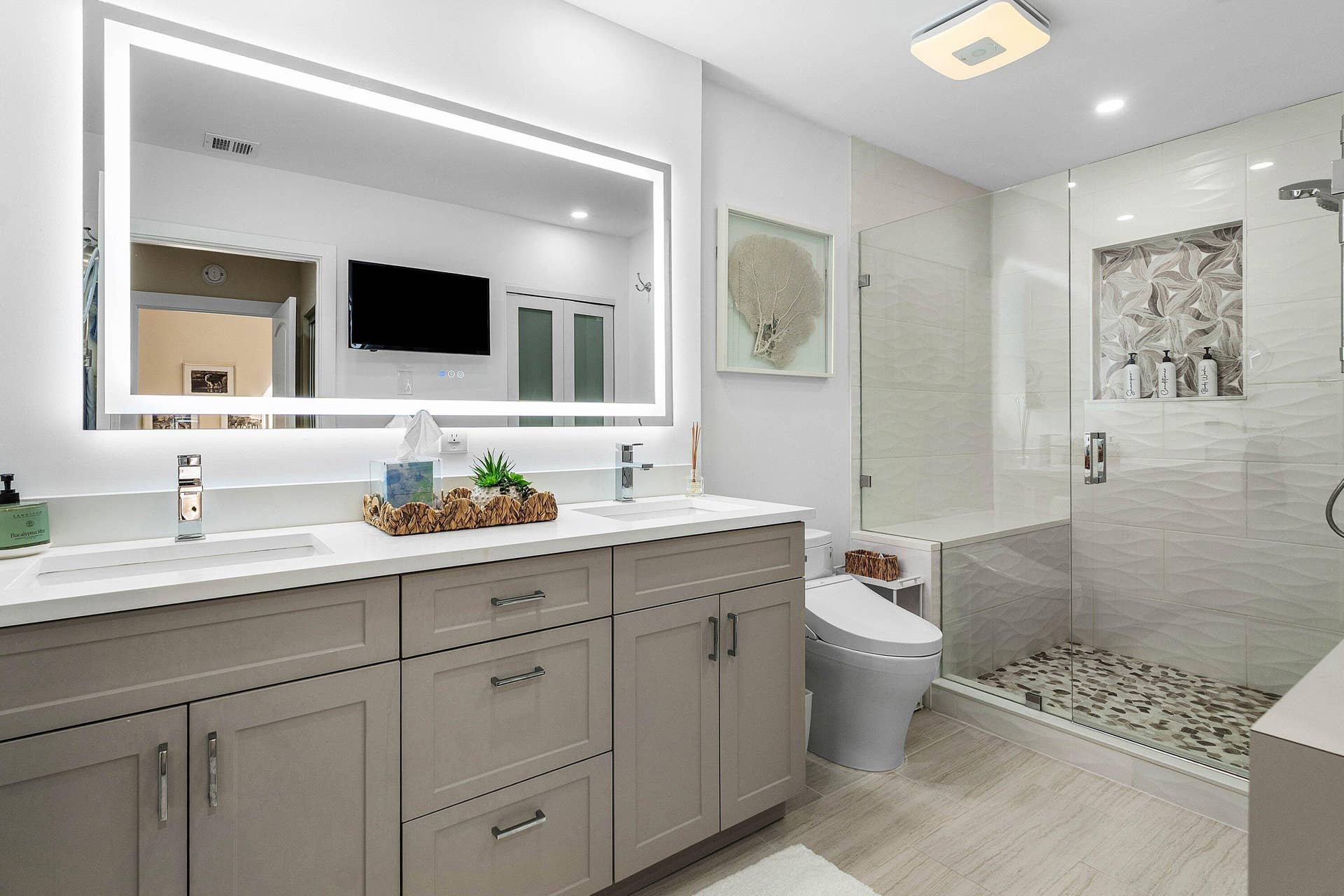 Dual-vanity cabinet with quartz countertop, wall-to-wall LED-backlit mirror, glass-enclosed shower with decorative tile inlay, and stone-tiled shower flooring