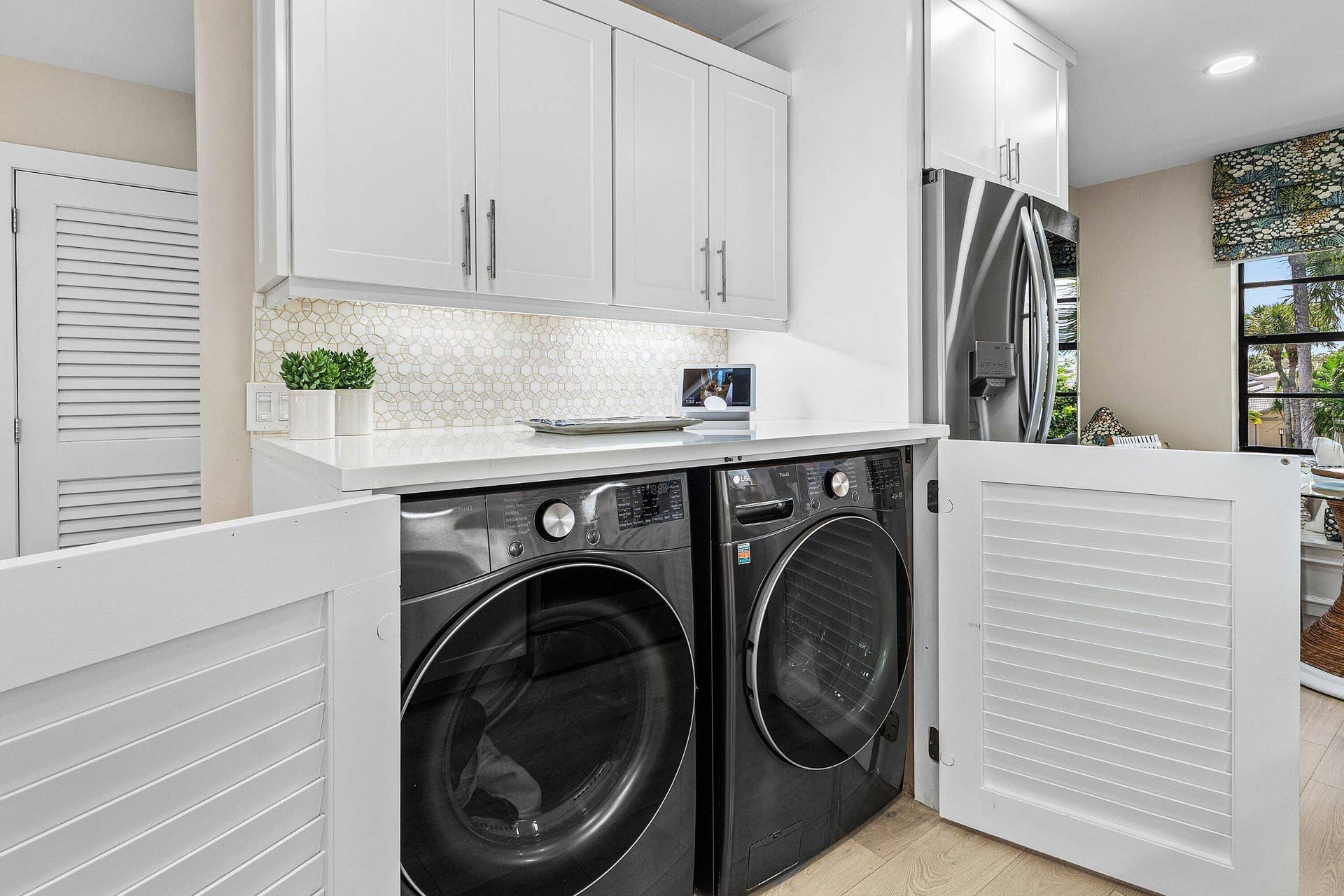 Laundry area featuring white cabinetry with brushed-metal pulls, stone-composite countertop, patterned hexagonal tile backsplash, and louvered storage doors