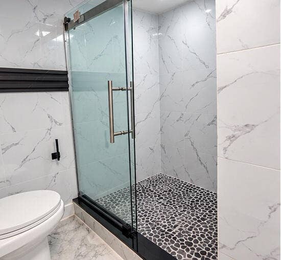 3601 SW 54th Avenue, West Park, FL 33023 Walk-in shower featuring a frameless glass enclosure, pebble-stone flooring, large-format veined tile surround, and matte black hardware