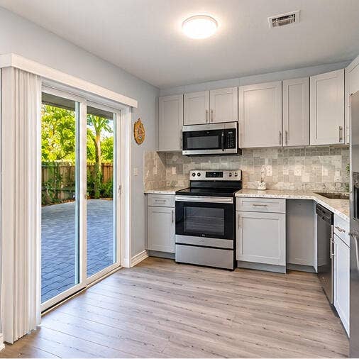 3601 SW 54th Avenue, West Park, FL 33023 Kitchen featuring shaker-style cabinetry, stainless steel appliances, stone-look countertops, patterned tile backsplash, and wood-finish flooring with sliding glass door access to a paved patio
