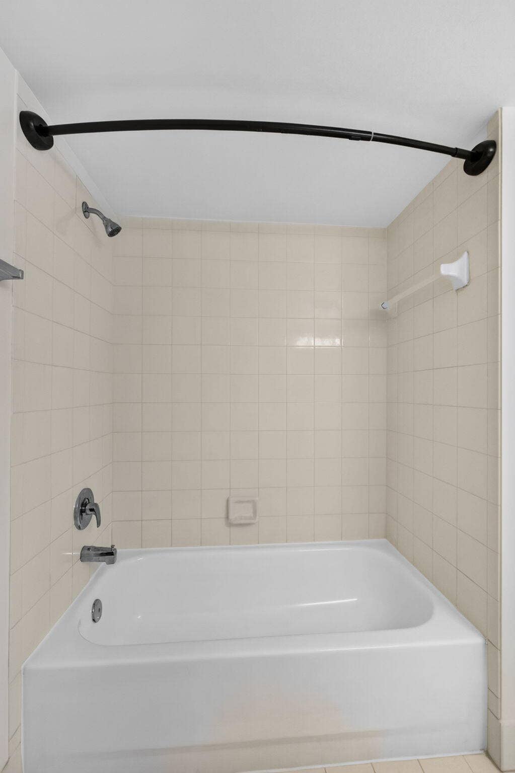 Bathtub with a white acrylic surround, ceramic wall tiling, polished chrome faucet, and a matte black curved shower rod
