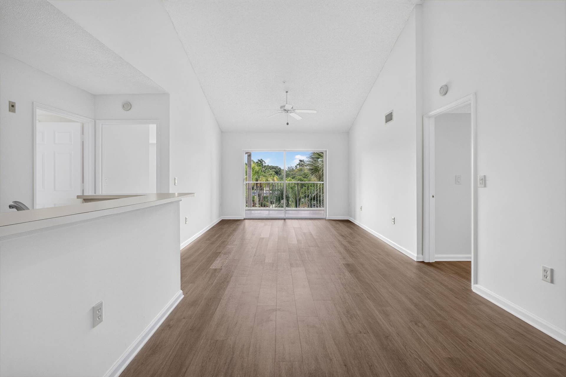 Vaulted ceilings featuring a central ceiling fan, vinyl plank flooring, a pass-through bar counter, and sliding glass doors with balcony access.