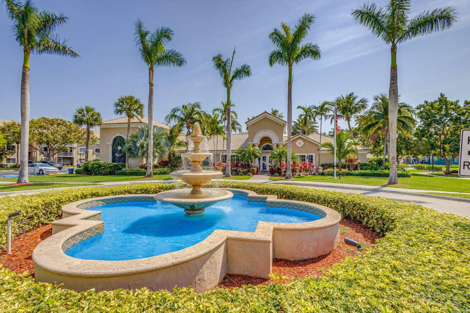 Tiered stone fountain centered within a manicured landscape featuring mature palm trees, sculpted hedges, and concrete walkways