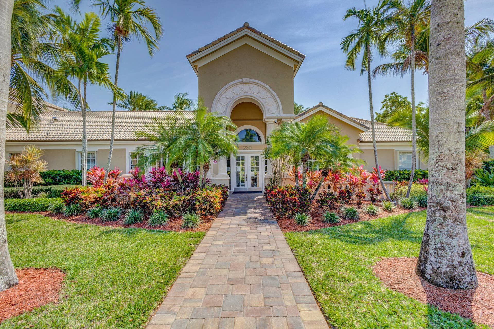 Exterior facade featuring a central arched entryway with decorative moulding, tile roof, and a paved stone walkway surrounded by mature palms and established tropical landscaping
