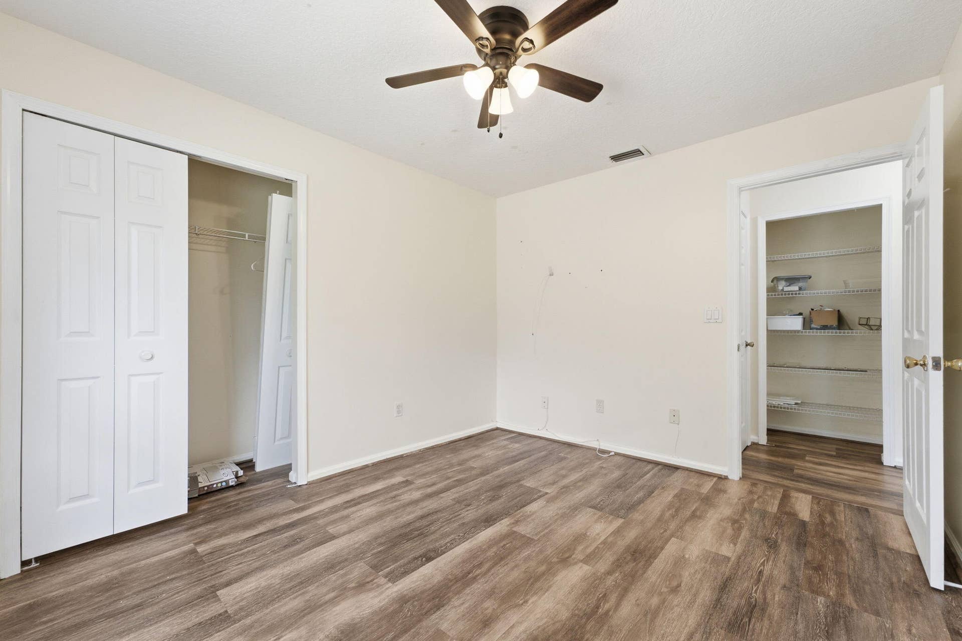 Room featuring wood-finish flooring, a ceiling fan with three-light fixture, dual-panel closet doors, and an open doorway revealing built-in wire shelving