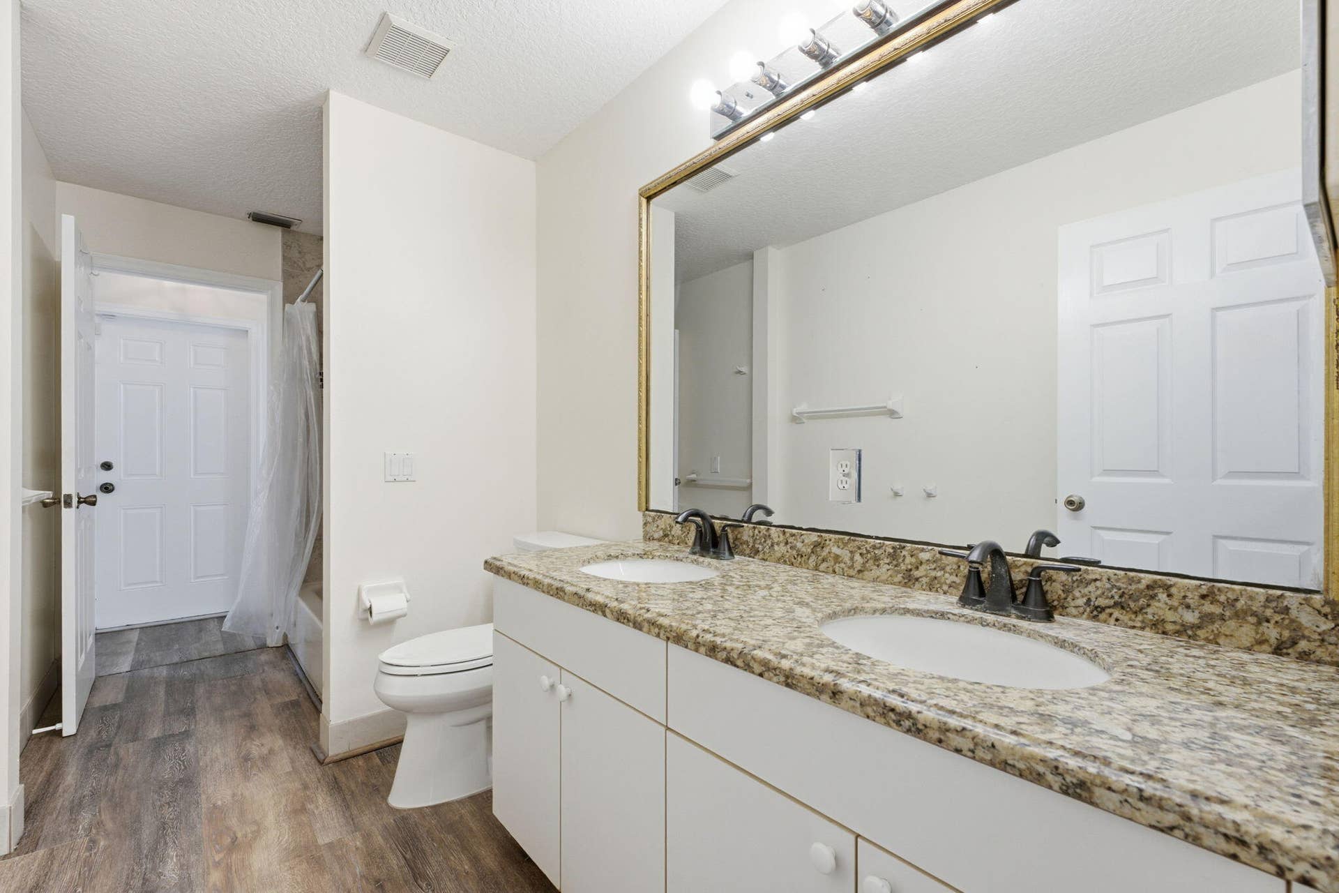 Dual-sink vanity with stone-finish countertop, undermount sinks, bronze-tone fixtures, wood-finish flooring, and large wall-mounted mirror