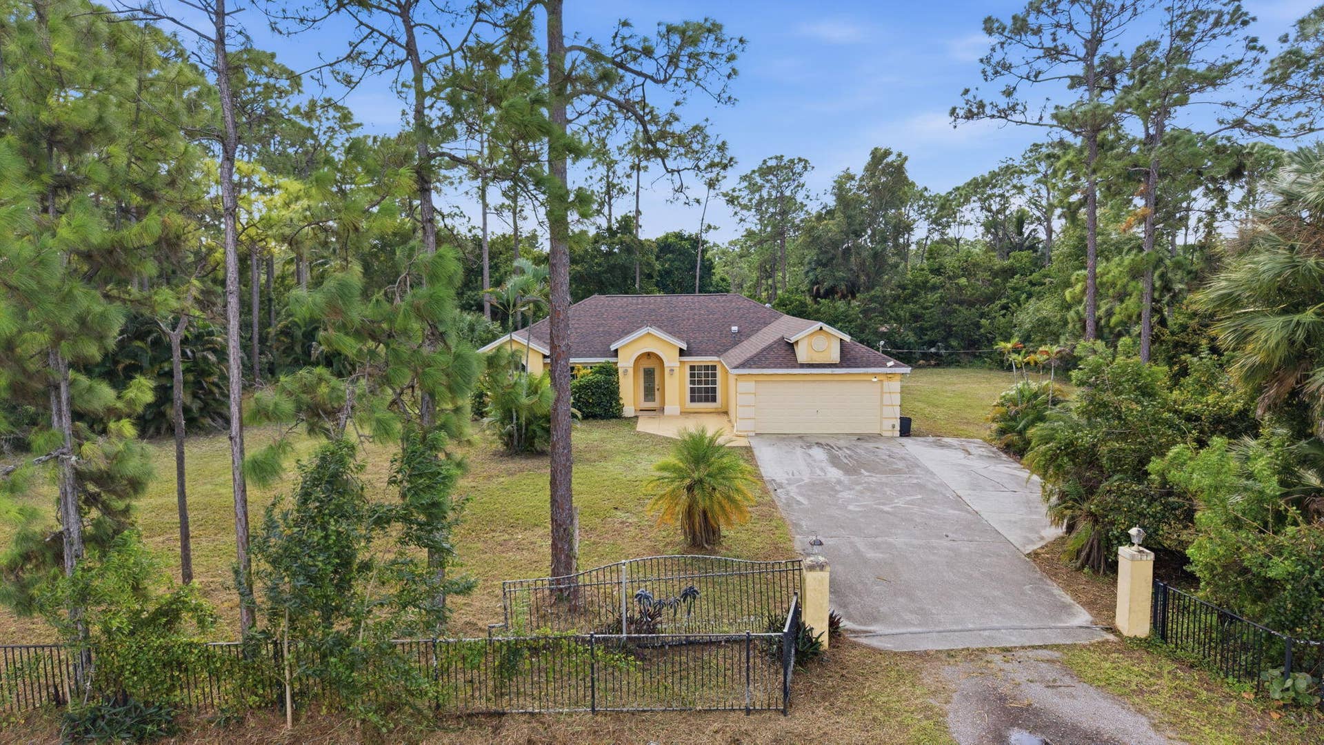 Single-story residence featuring a hipped roof, attached garage, concrete driveway, and perimeter metal fencing set among mature pine trees